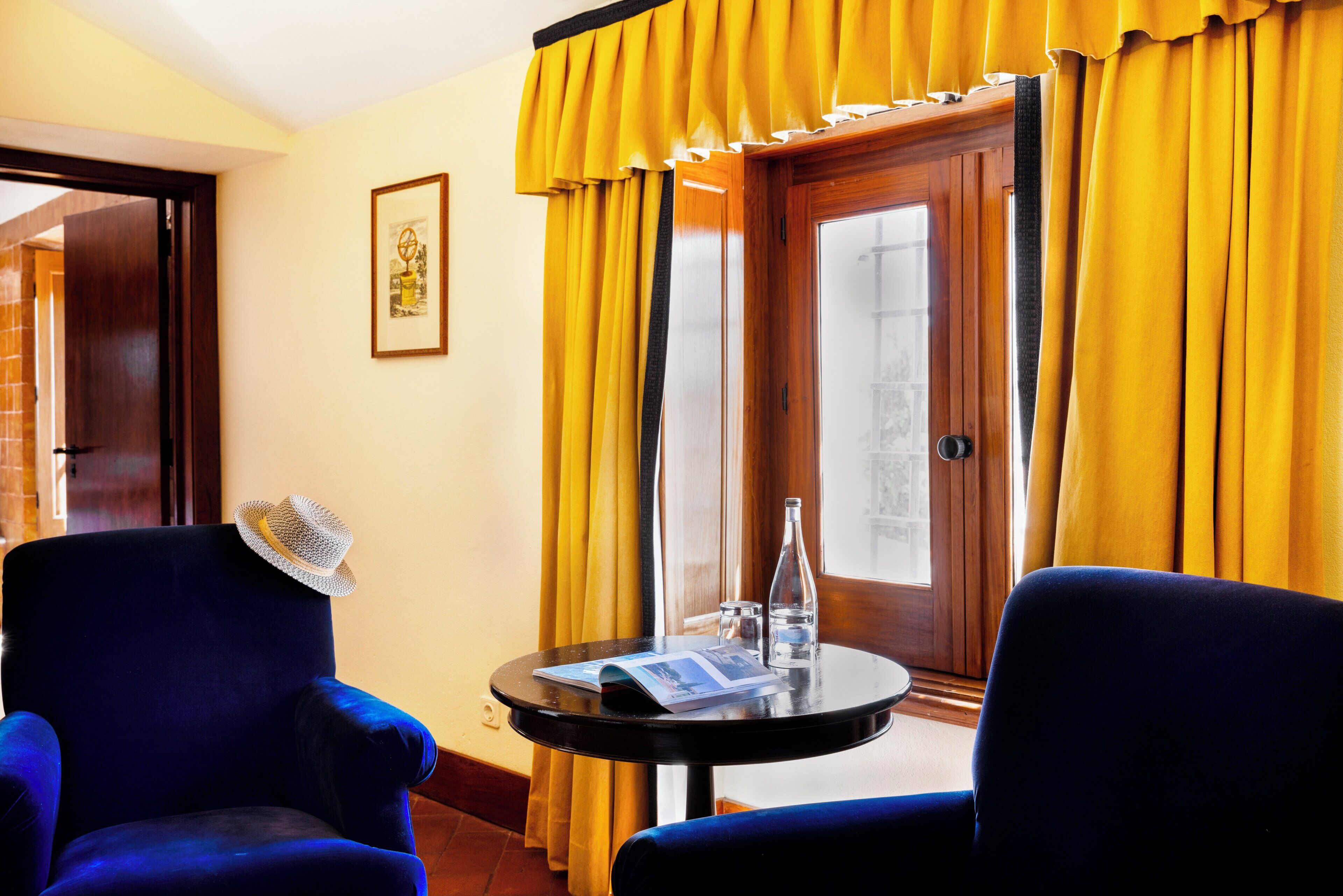 Une pièce avec deux fauteuils bleus, une petite table ronde sur laquelle se trouvent une bouteille et un menu, et une porte ornée de rideaux jaunes.