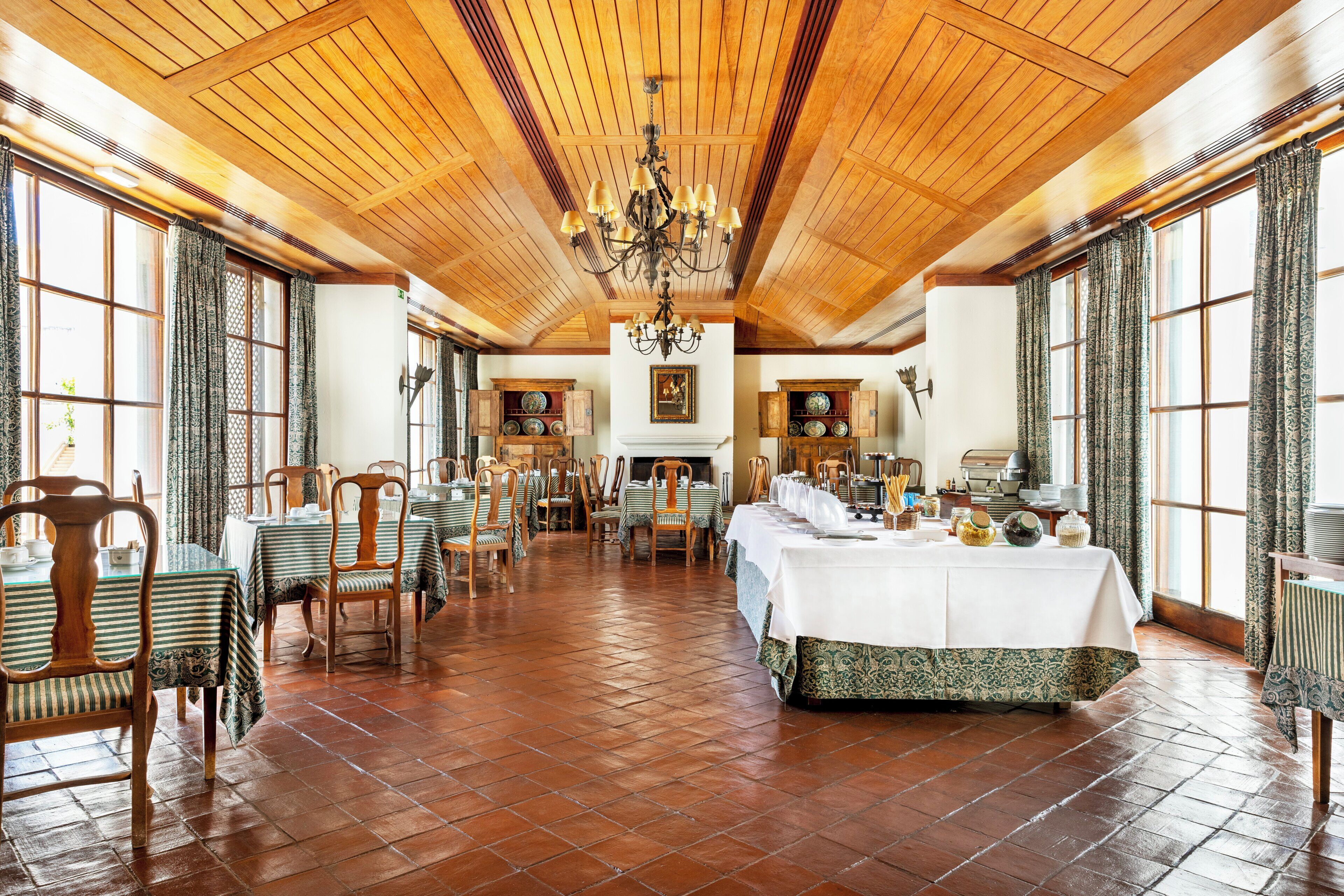 A spacious dining hall with high wooden ceilings, a central chandelier, and large windows with patterned curtains.