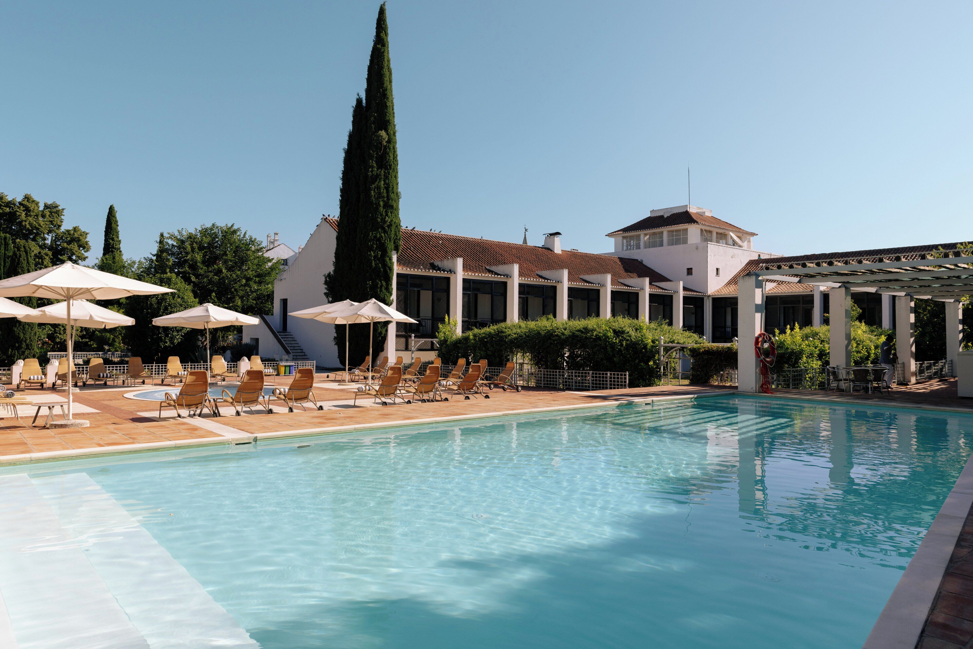 Piscina con sillas de descanso y sombrillas, rodeada por un edificio con techo de tejas y un árbol alto.
