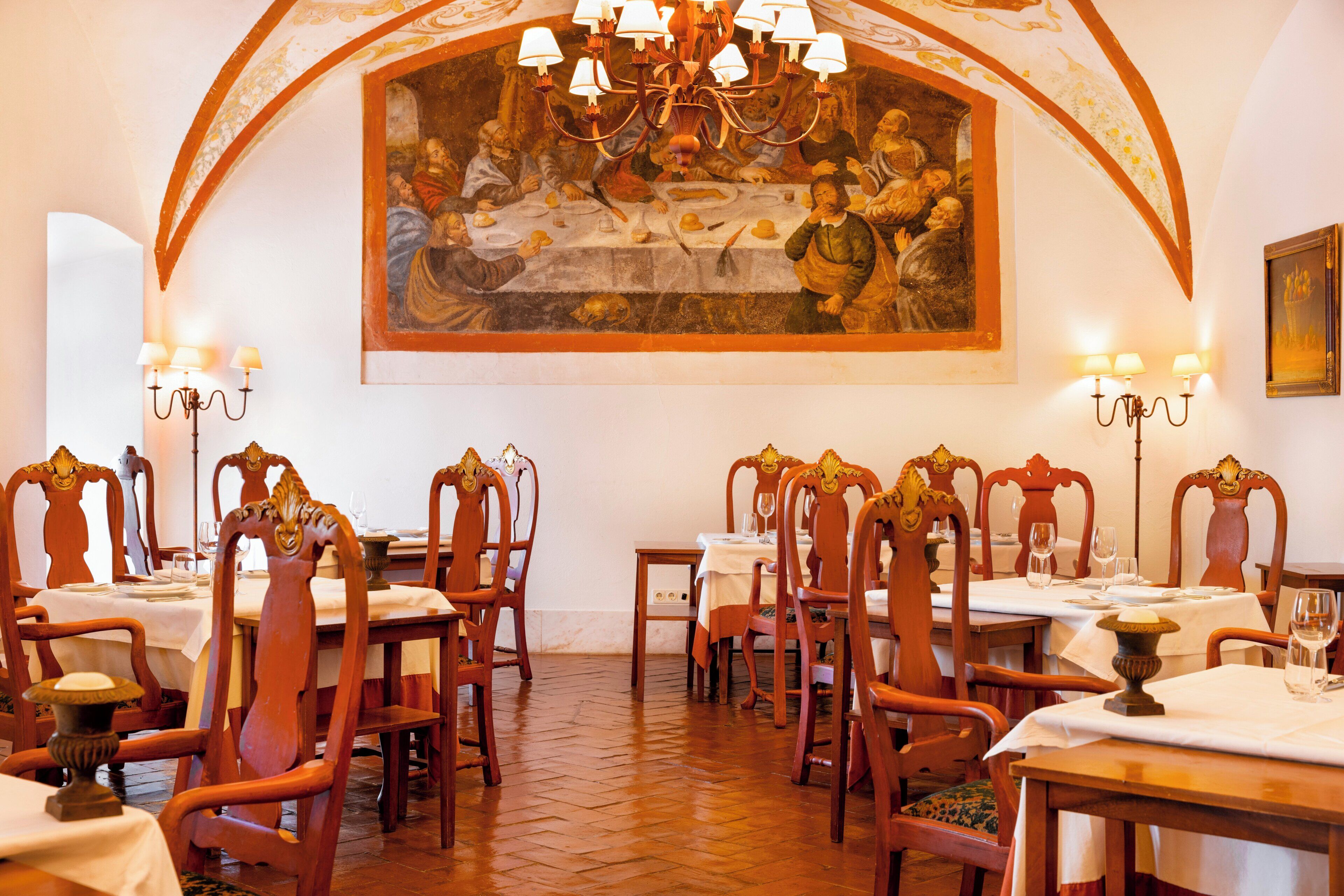 Un comedor con sillas de madera ornamentadas, mesas cubiertas con manteles blancos y un cuadro grande sobre las mesas que representa una escena histórica.