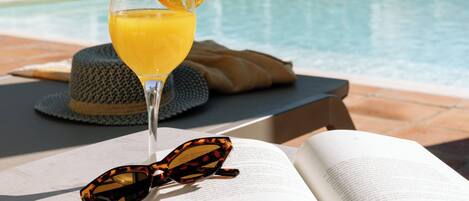 Escena junto a la piscina con una bebida, gafas de sol y un libro abierto.