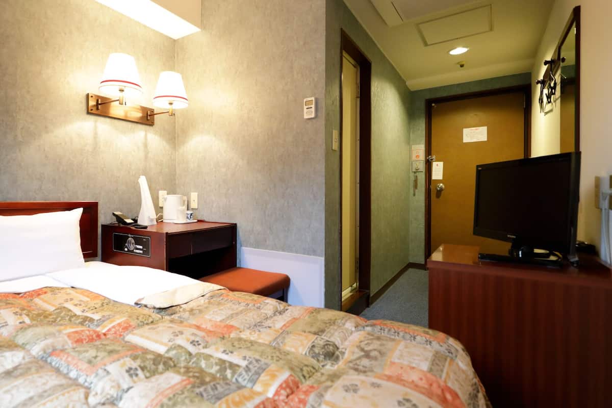 A hotel room with a bed, bedside table, lamp, TV, and a door leading to another room.