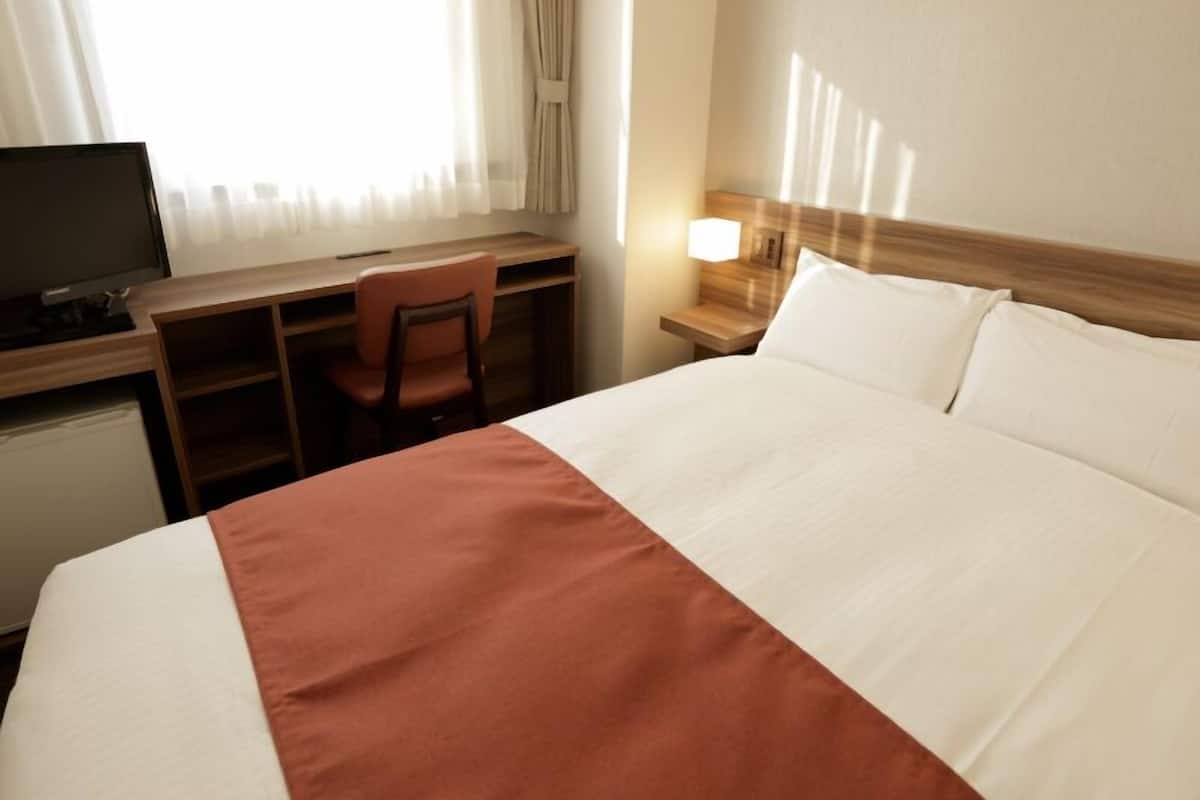 A hotel room with a bed, a desk with a chair, a television, and a nightstand with a lamp.