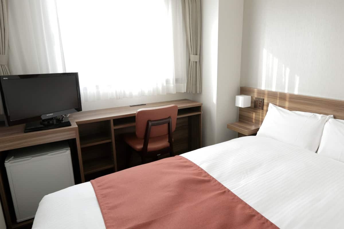 A hotel room with a bed, a desk with a computer, a chair, a nightstand, and a small refrigerator.