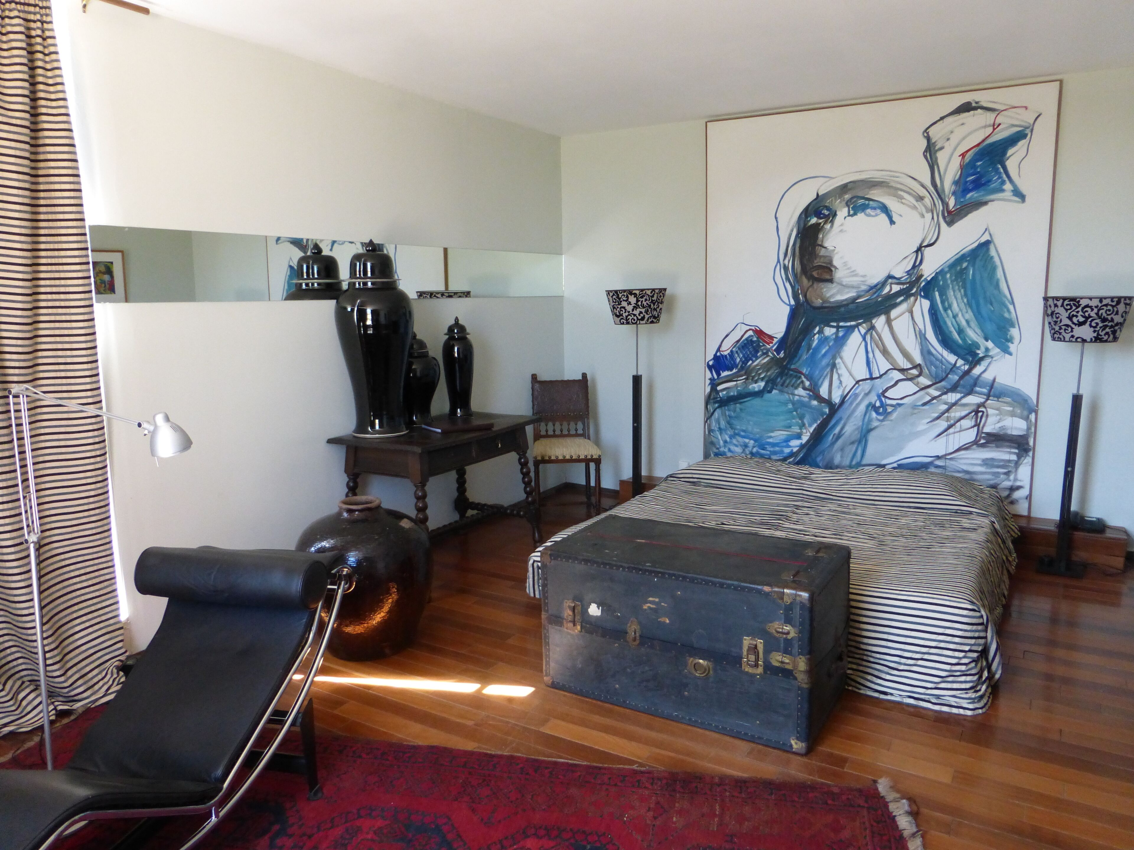 A bedroom with a large bed, a desk, a chair, a suitcase, and a painting on the wall.