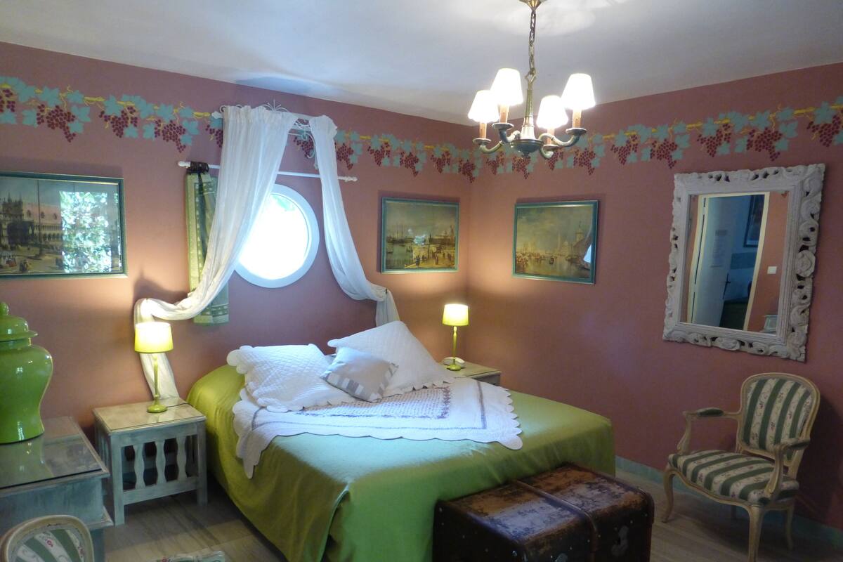 Une chambre à coucher comprenant un lit, deux lampes de chevet, une table de chevet, une chaise, un miroir et une fenêtre avec des rideaux.