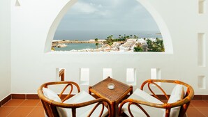 Un balcón con sillas de mimbre y una mesa, con vista a un pueblo costero con edificios y palmeras.