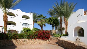 Un edificio blanco con ventanas y balcones arqueados, rodeado de palmeras y arbustos florecientes de color rojo.