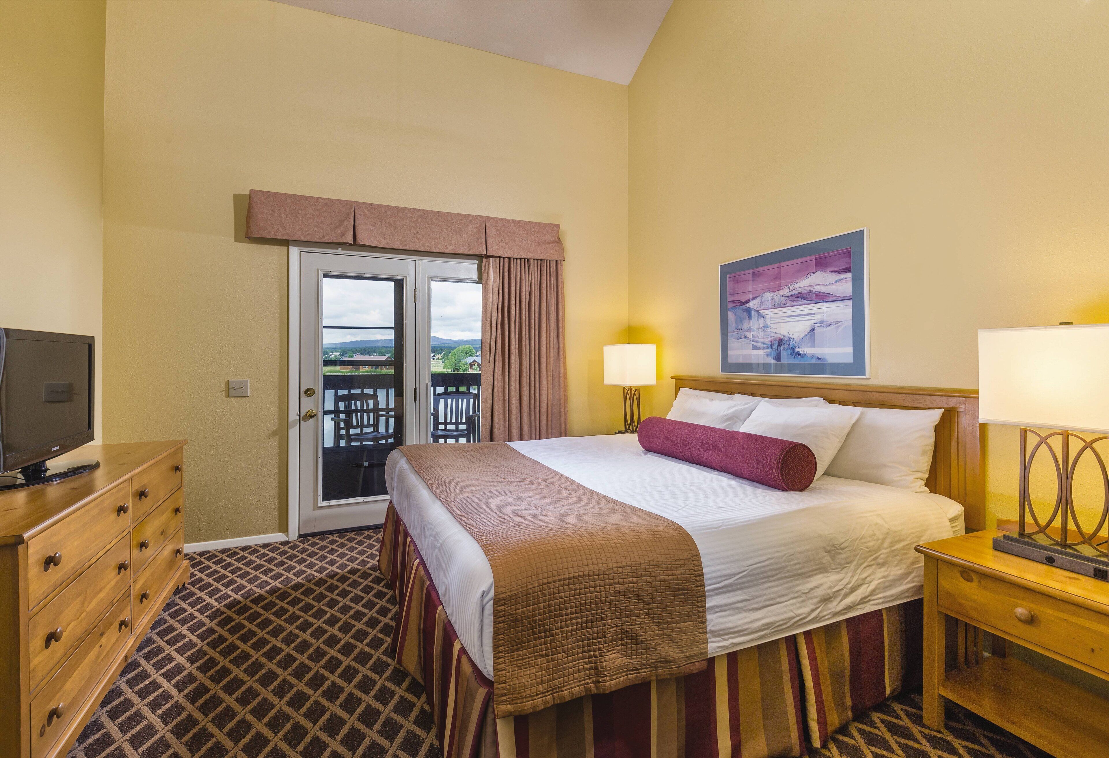 Une chambre d’hôtel avec un lit, un téléviseur posé sur une commode, une table de chevet avec une lampe, et une porte donnant sur un espace extérieur.