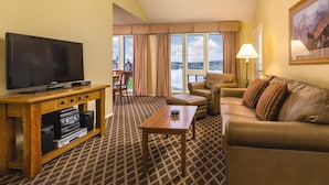 Habitación de hotel con un televisor de pantalla plana grande, sofá de cuero, sillón y una mesita. Se ve una ventana con vistas a un lago y casas.