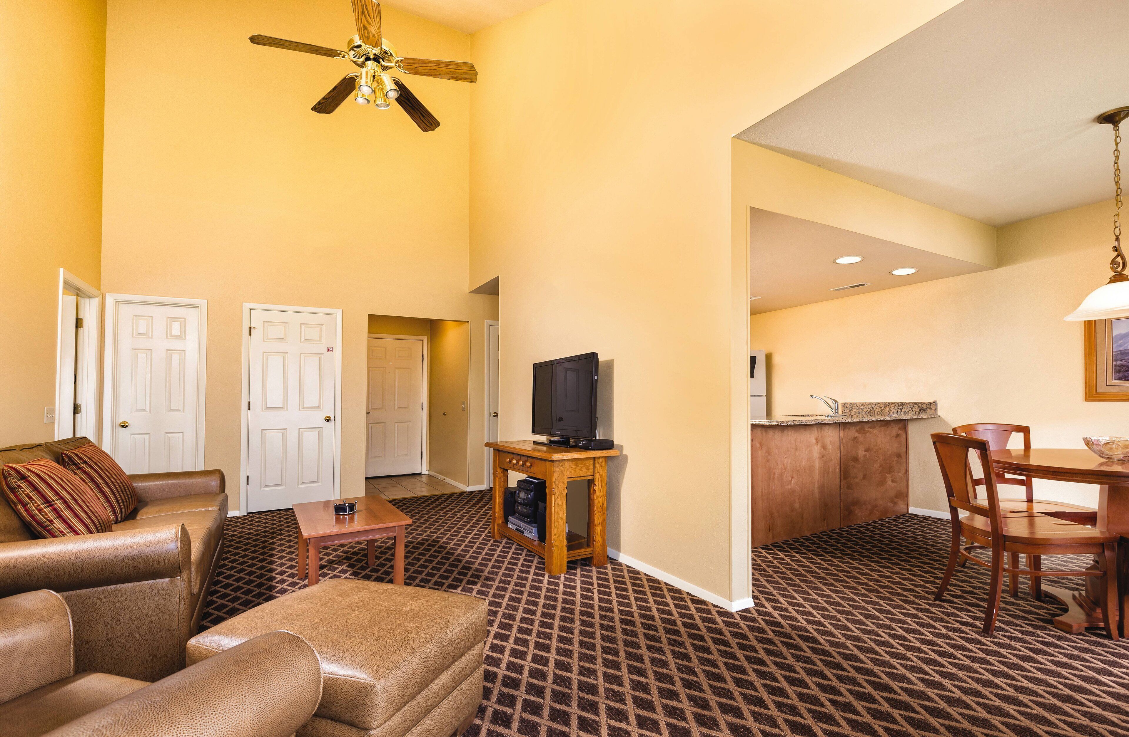 Habitación de hotel con sala de estar que incluye un sofá, una mesa de centro y un televisor sobre un soporte de madera. Cuenta con una mesa de comedor con sillas y una cocina con lavamanos y encimera.