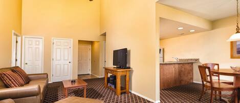 Habitación de hotel con sala de estar que incluye un sofá, una mesa de centro y un televisor sobre un soporte de madera. Cuenta con una mesa de comedor con sillas y una cocina con lavamanos y encimera.