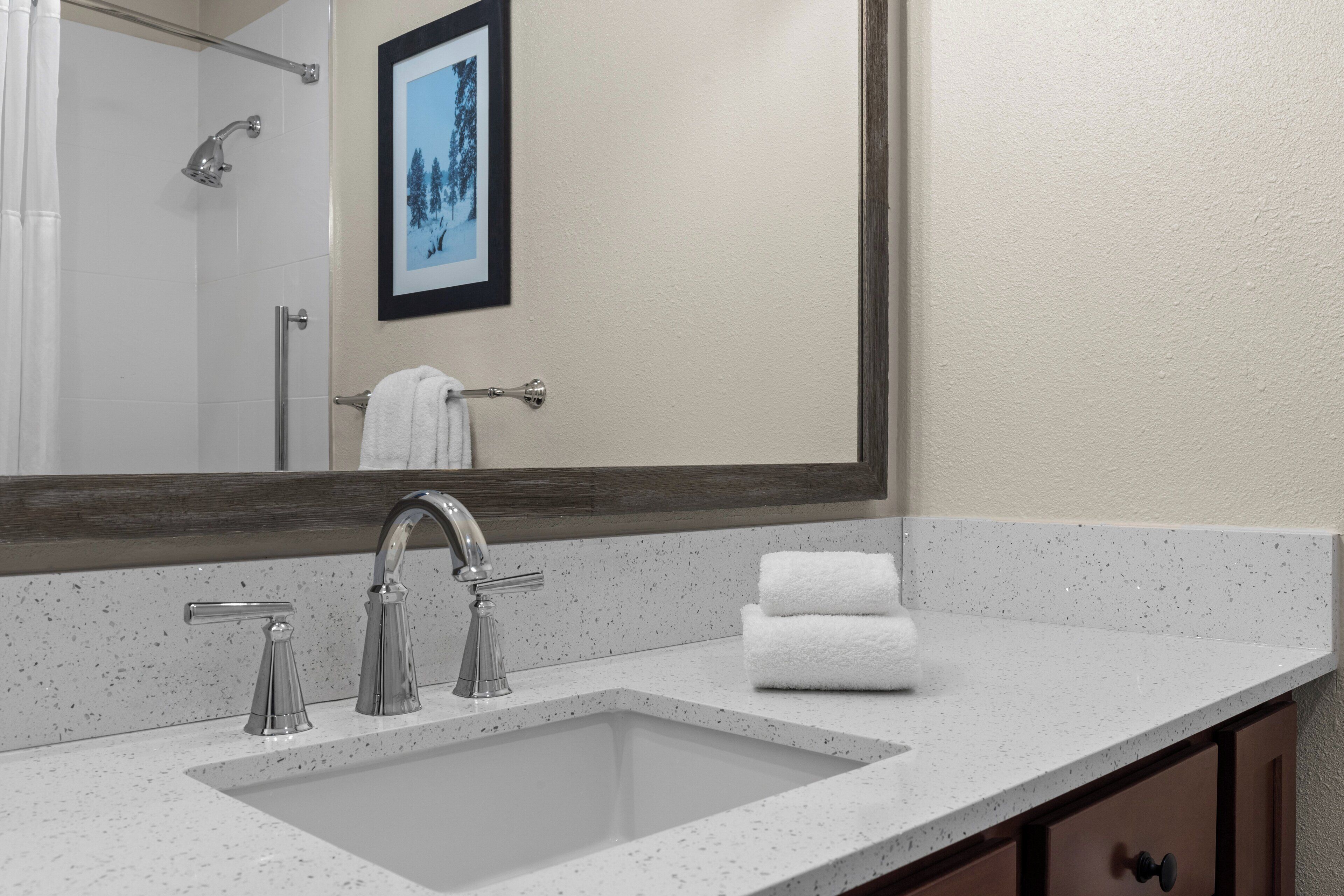 A bathroom with a large mirror, a sink with two faucets, a towel rack with a towel, and a framed picture on the wall.