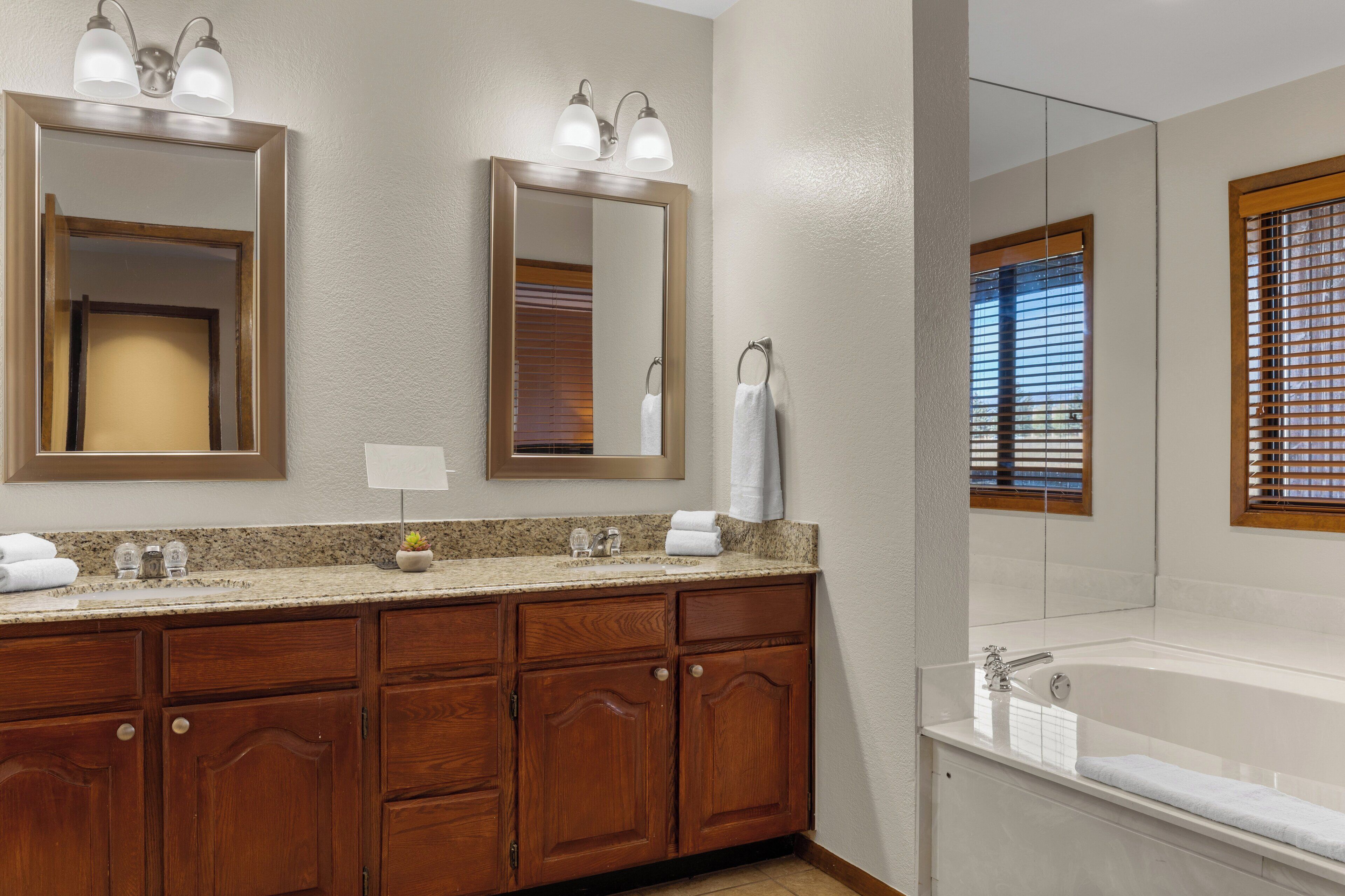 A bathroom with a wooden vanity, a large mirror, a bathtub, and a window with blinds.