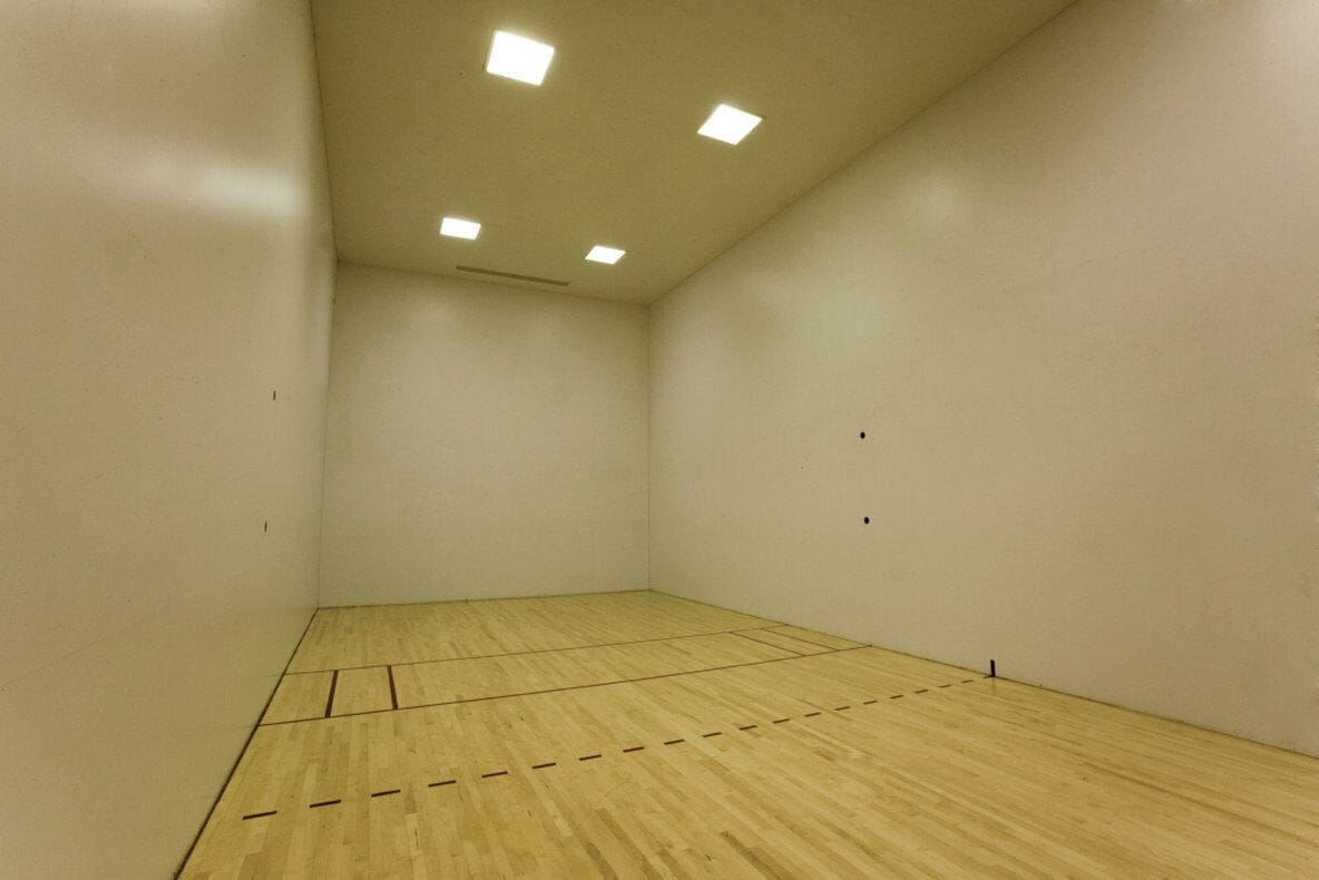 Un court de squash doté d’un plancher en bois et de murs blancs, et équipé de luminaires au plafond.