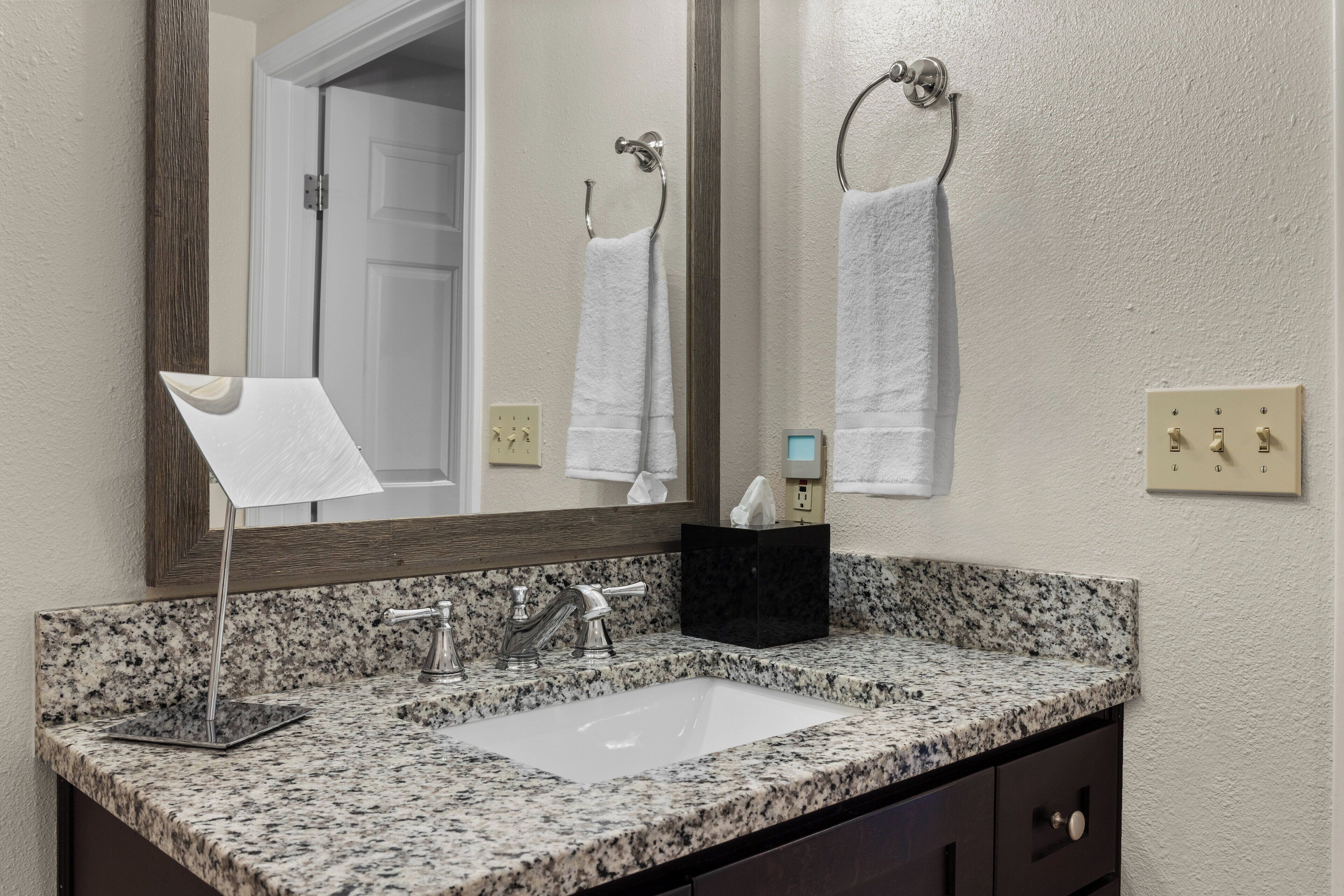 A bathroom with a granite countertop sink, a mirror, a towel rack, and a black soap dispenser.