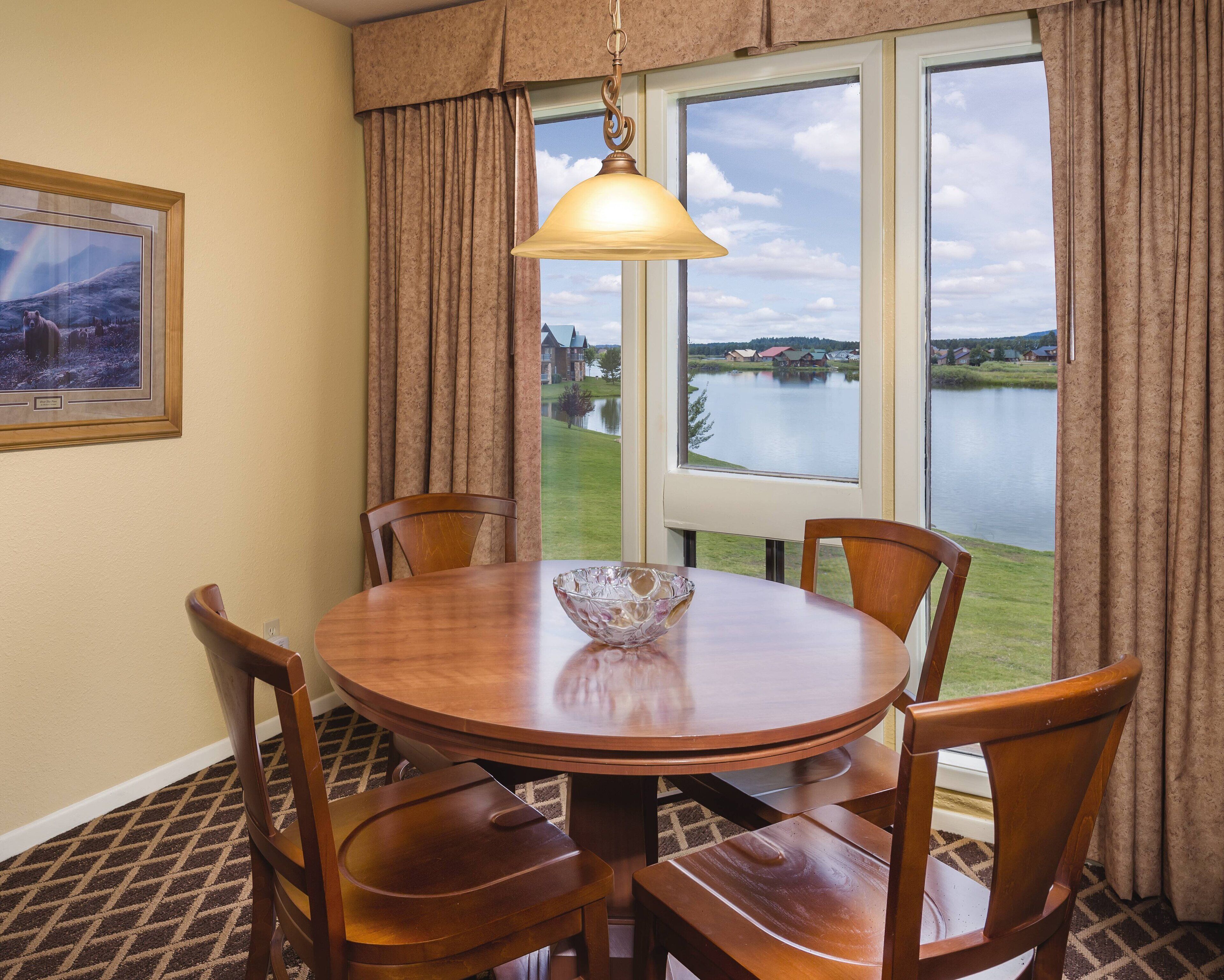 Comedor con mesa redonda de madera, cuatro sillas, lámpara colgante y ventana con vista a un lago y edificios.