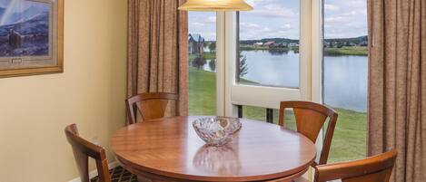Comedor con mesa redonda de madera, cuatro sillas, lámpara colgante y ventana con vista a un lago y edificios.