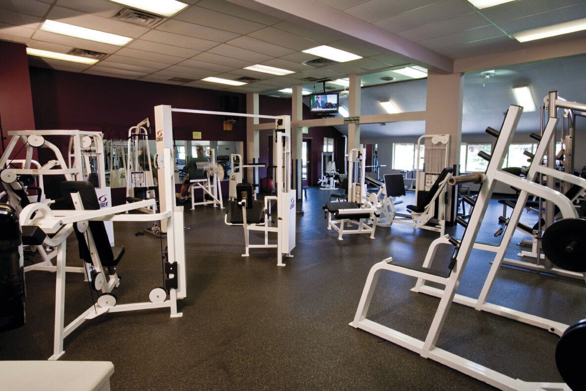 Et fitnesscenter med diverse træningsmaskiner, et fjernsyn på væggen og store vinduer, der lukker naturligt lys ind.
