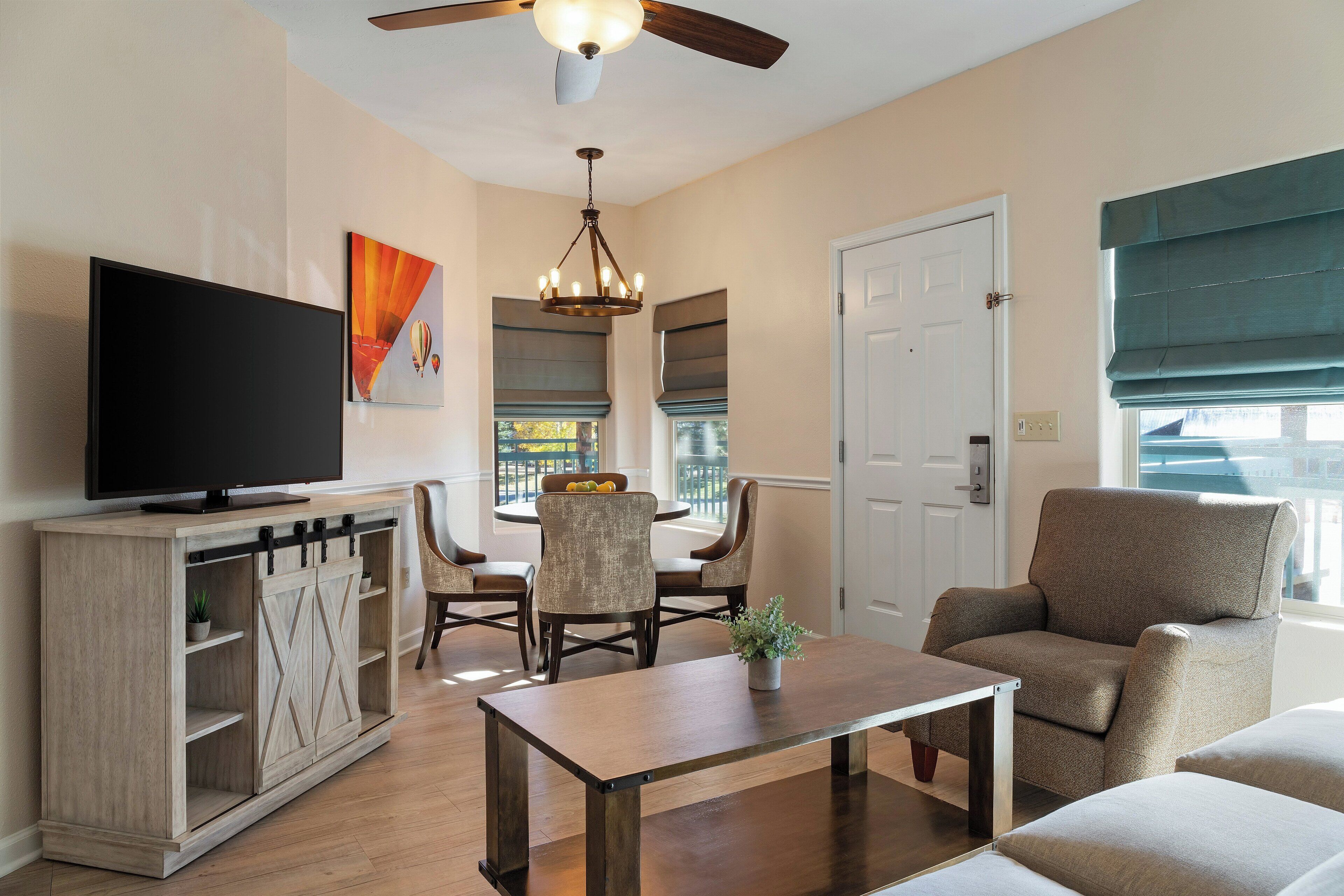 A living room with a TV, a dining table, and chairs. There is a ceiling fan, a chandelier, and a window with blinds.
