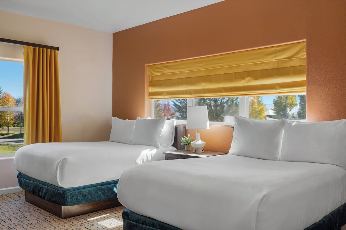 A hotel room with two beds, a window with a yellow blind, a bedside table with a lamp, and a view of trees outside.