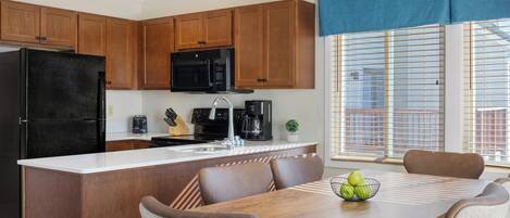 Una cucina moderna con mobili in legno, un frigorifero nero, un forno a microonde e una macchina da caffè. C'è un tavolo da pranzo con sedie e una ciotola di frutta.