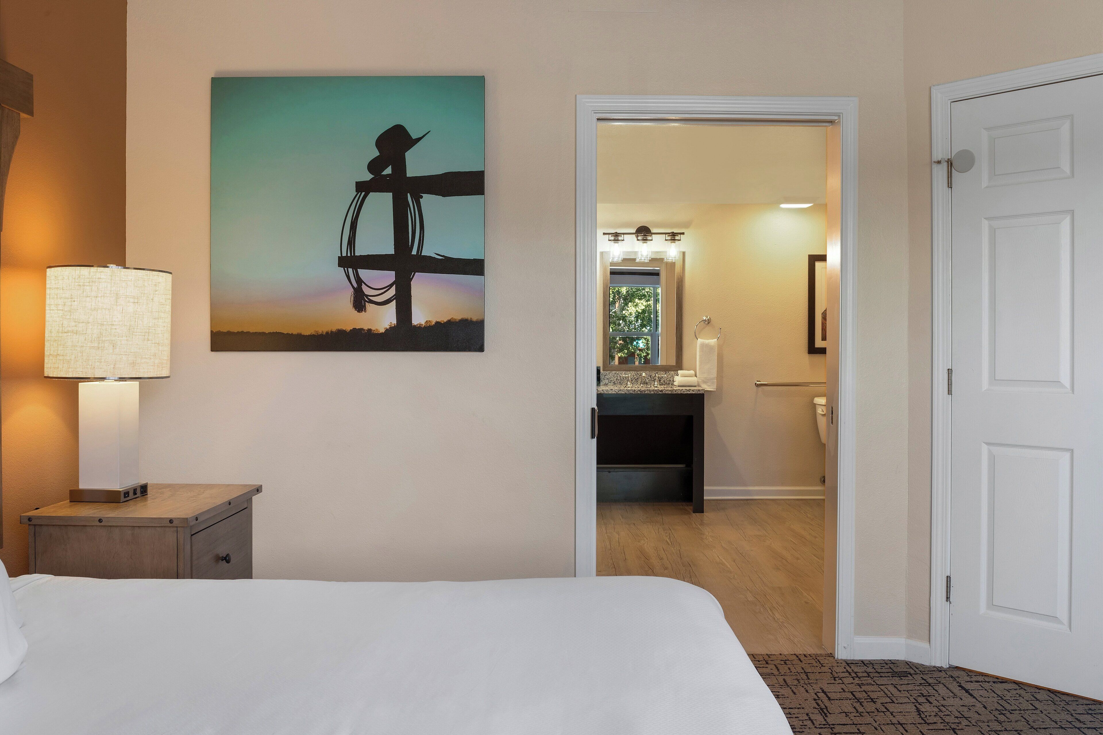 A hotel room with a bed, a nightstand with a lamp, a painting of a cowboy on a horse, and a bathroom visible through an open door.