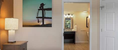 A hotel room with a bed, a nightstand with a lamp, a painting of a cowboy on a horse, and a bathroom visible through an open door.