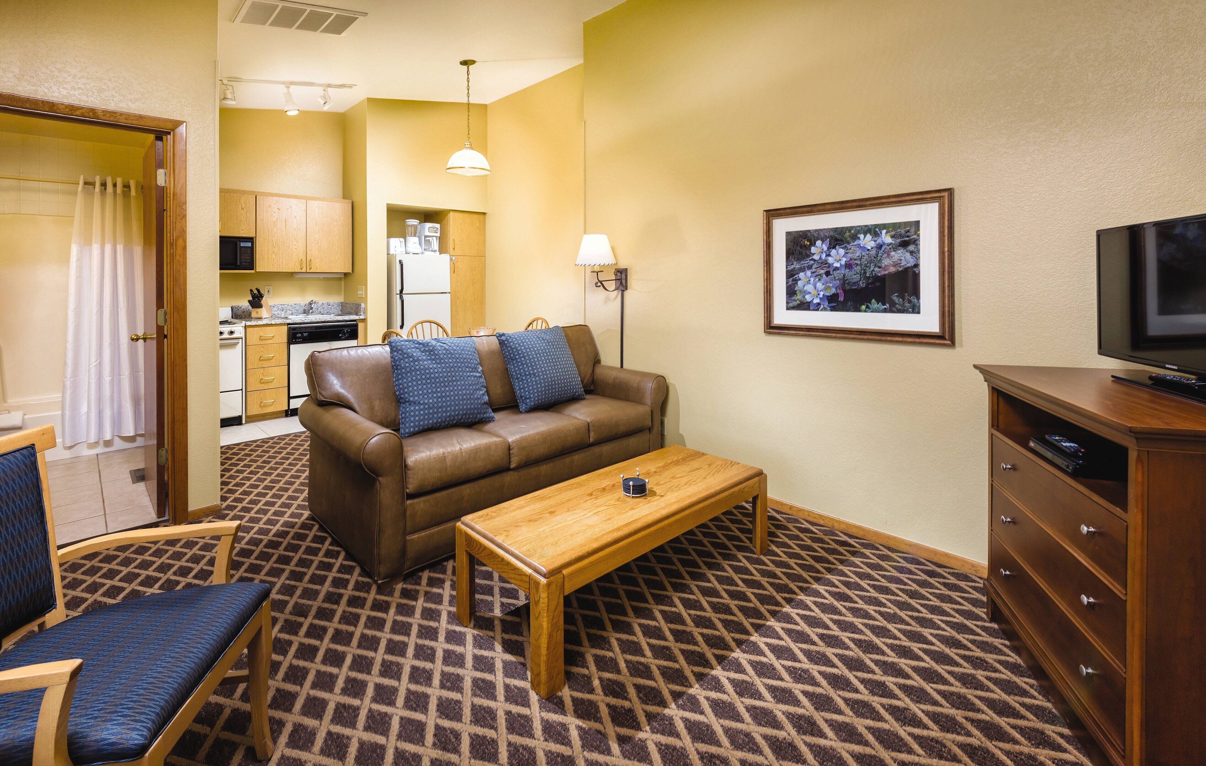 A hotel room with a brown leather sofa, a wooden coffee table, a TV, a dresser, and a kitchen area in the background.