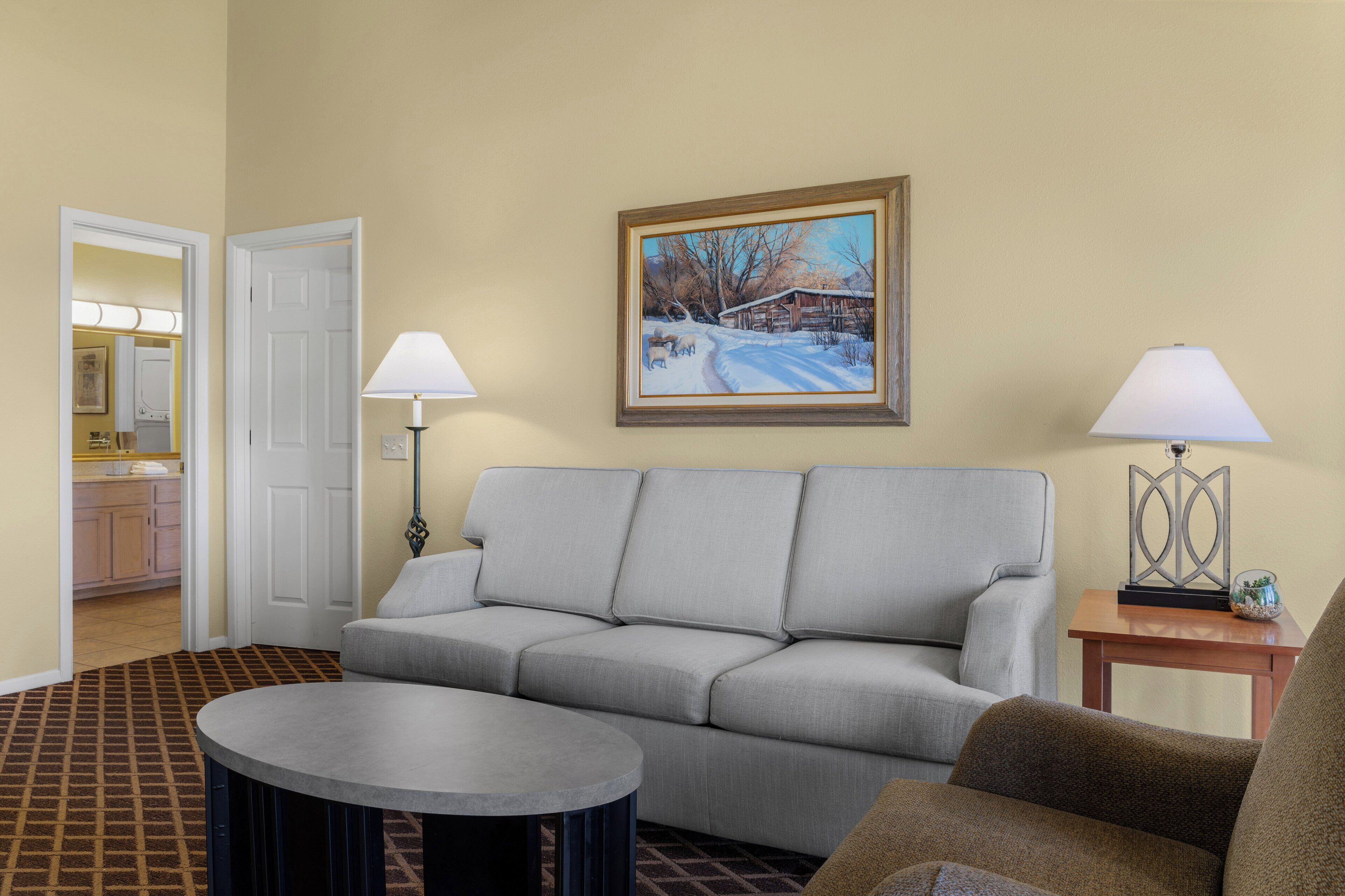 A living room with a gray sofa, a round coffee table, two lamps, and a painting of a snowy landscape.