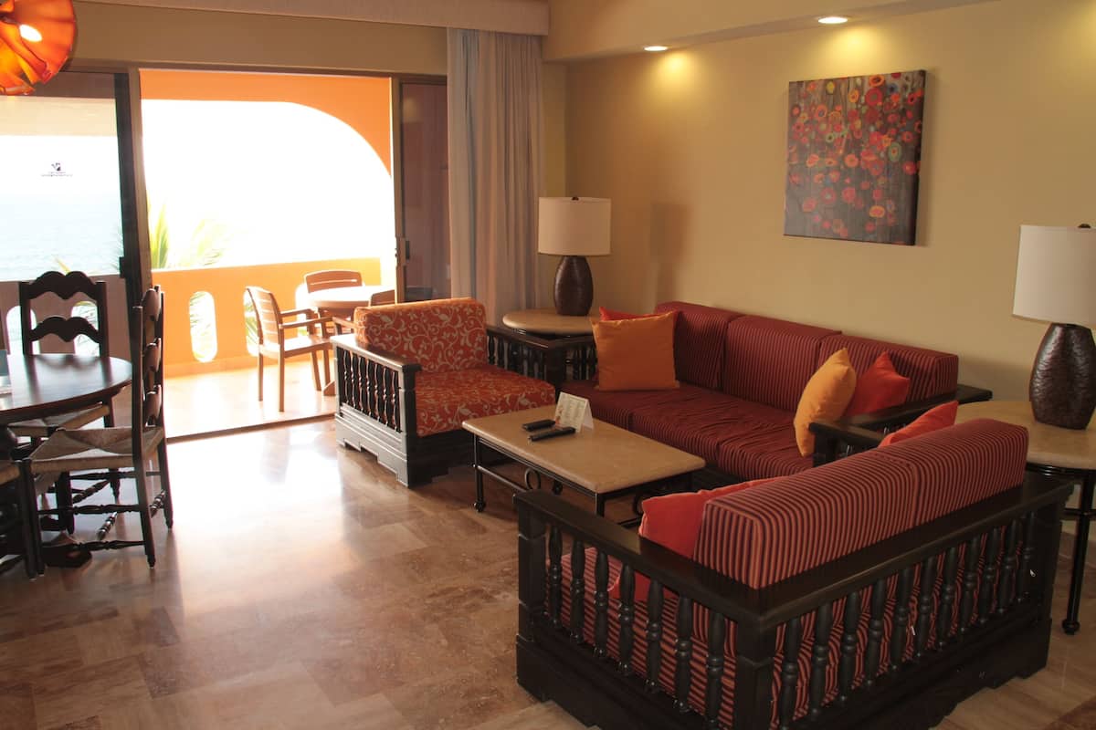 A hotel room with a sofa set, a dining table with chairs, a small table with a lamp, and a painting on the wall.