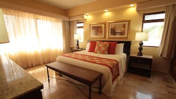 Una habitación de hotel con una cama grande, un banco, dos mesitas de noche con lámparas y una ventana con cortinas.