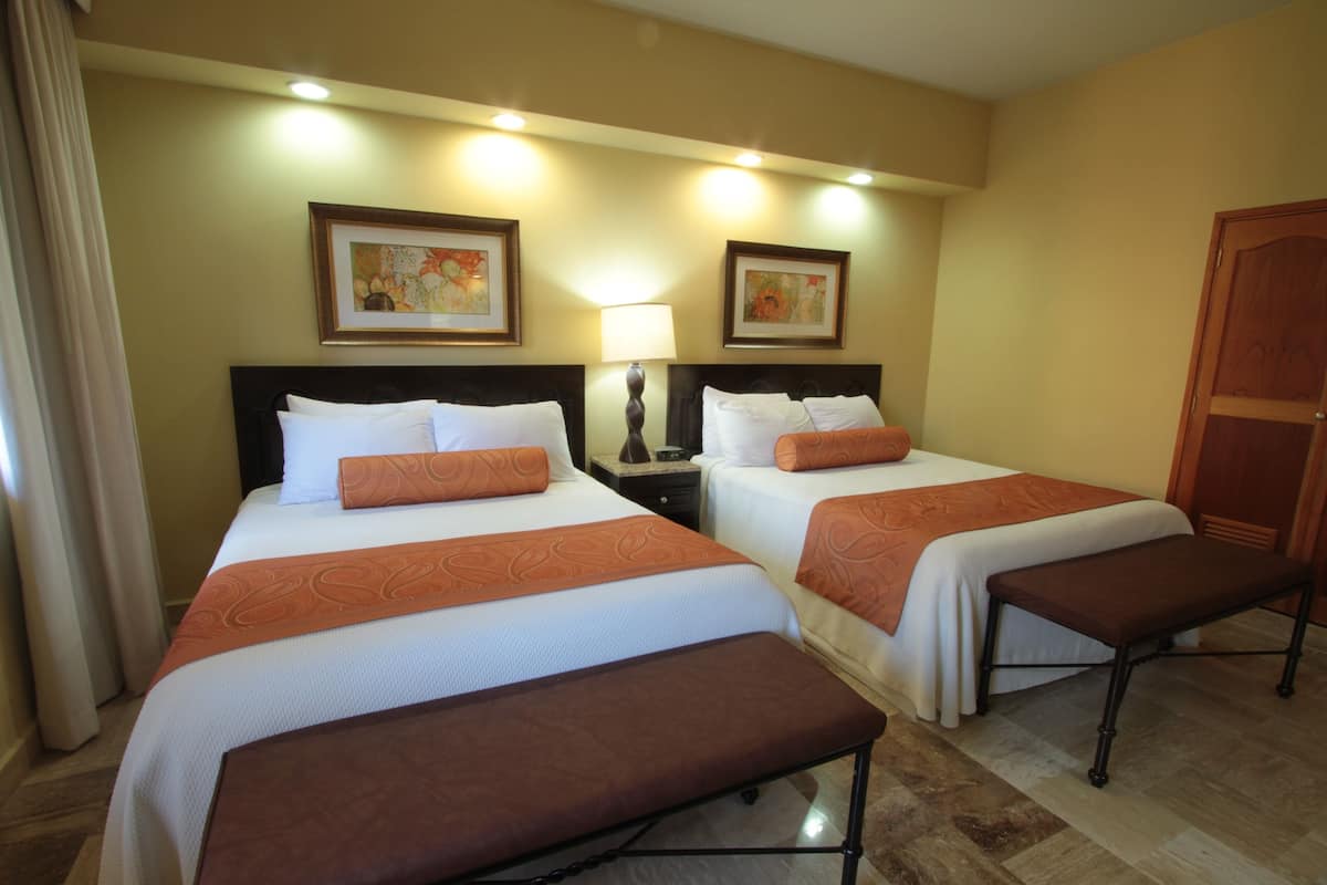 A hotel room with two beds, each with a brown bench, white bedding, and orange decorative pillows. There are framed artworks on the wall and a wooden door.