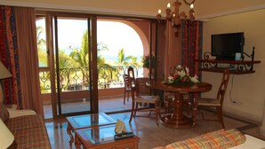 Een kamer met een bank, een eettafel, stoelen, een televisie en een balkon met uitzicht op palmbomen en de oceaan.