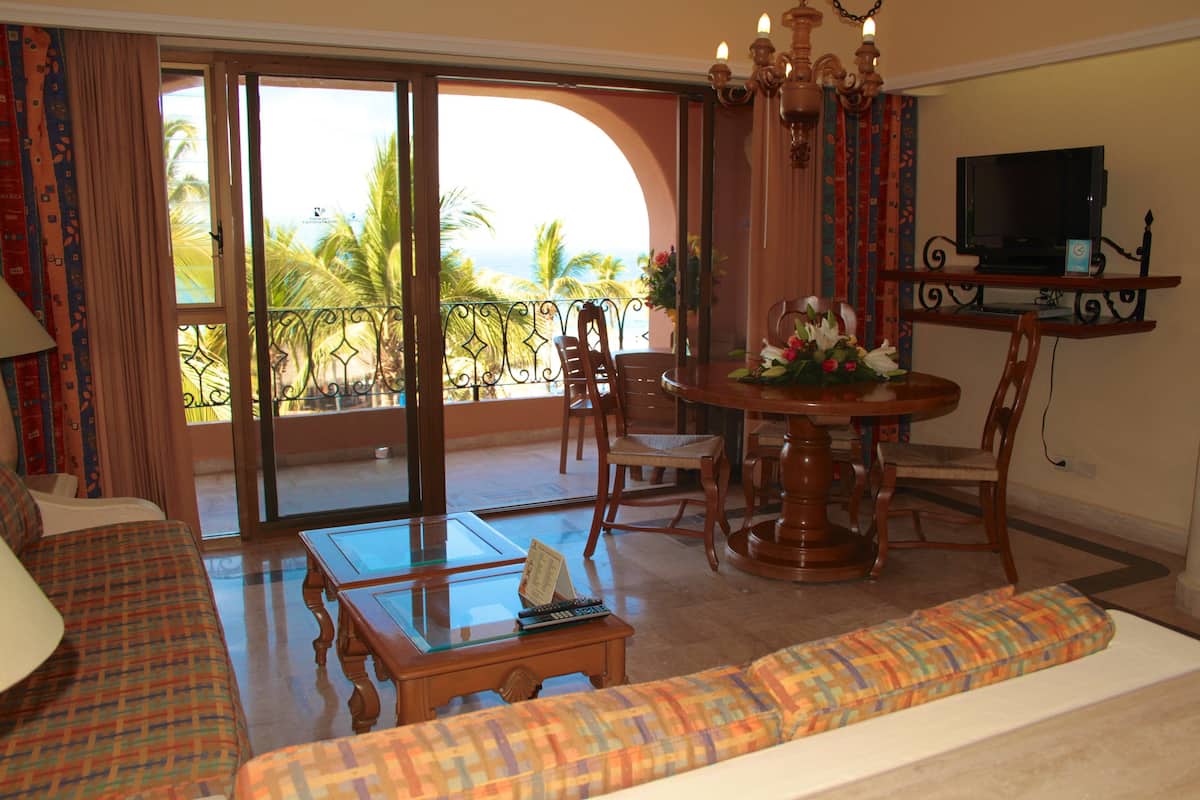 A room with a sofa, a dining table, chairs, a TV, and a balcony with a view of palm trees and the ocean.