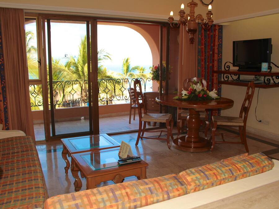 A room with a sofa, a dining table, chairs, a TV, and a balcony with a view of palm trees and the ocean.