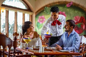 A restaurant interior with wooden furniture, a mural with floral designs, and a waiter serving a drink. - Holiday Inn Club Vacations The Royal Sands by IHG - All inclusive (Cancun)
