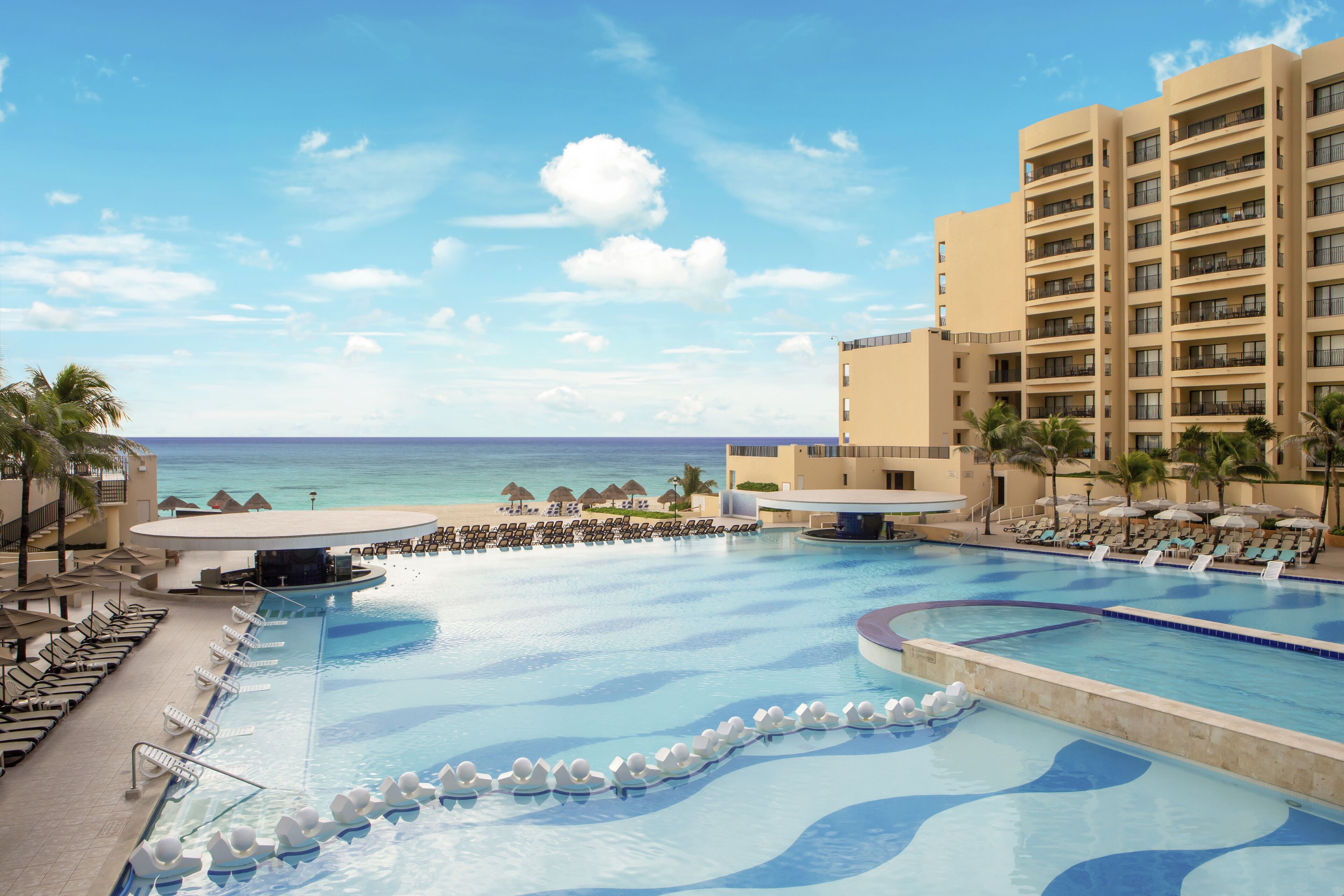 Una piscina de resort con sillones de descanso y sombrillas, vista a la playa y un edificio con balcones.