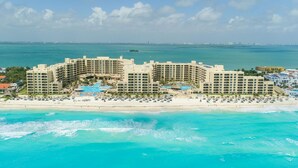 Un hotel frente a la playa con varias construcciones, una zona de piscina y una playa con sombrillas.
