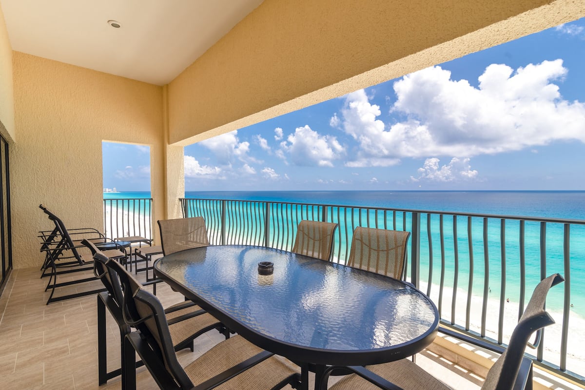 A balcony with a glass table and chairs, overlooking a beach with turquoise water and a clear sky.