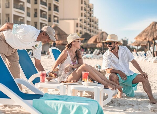 A beach scene with people relaxing on sun loungers, a table with drinks and snacks, and thatched umbrellas in the background. - Holiday Inn Club Vacations The Royal Sands by IHG - All inclusive (Cancun)