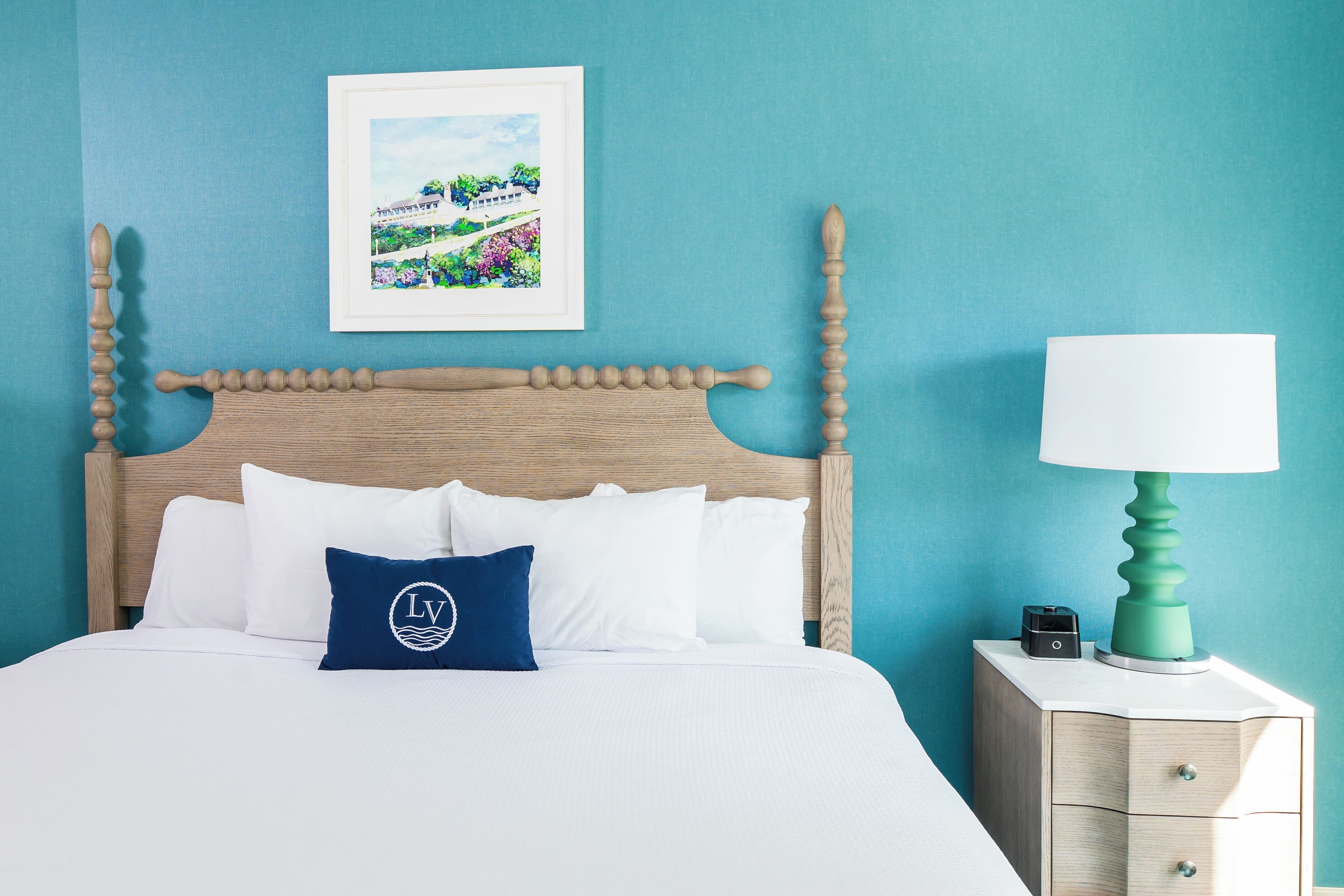 A neatly made bed with white linens and a single blue pillow, a wooden headboard, a framed picture on the wall, and a bedside table with a lamp and a small object.