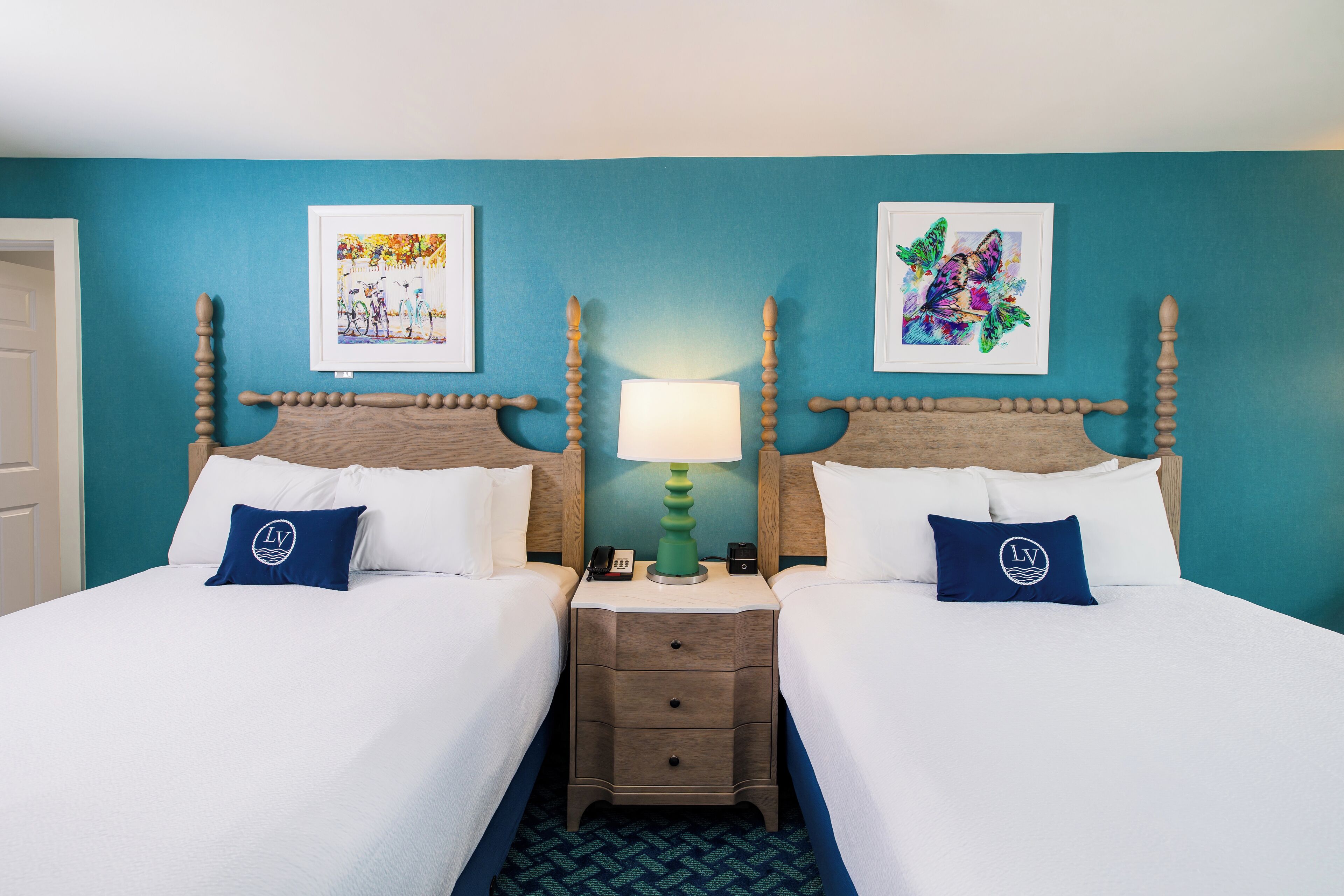 A hotel room with two beds, a nightstand with a lamp, and artwork on the walls.