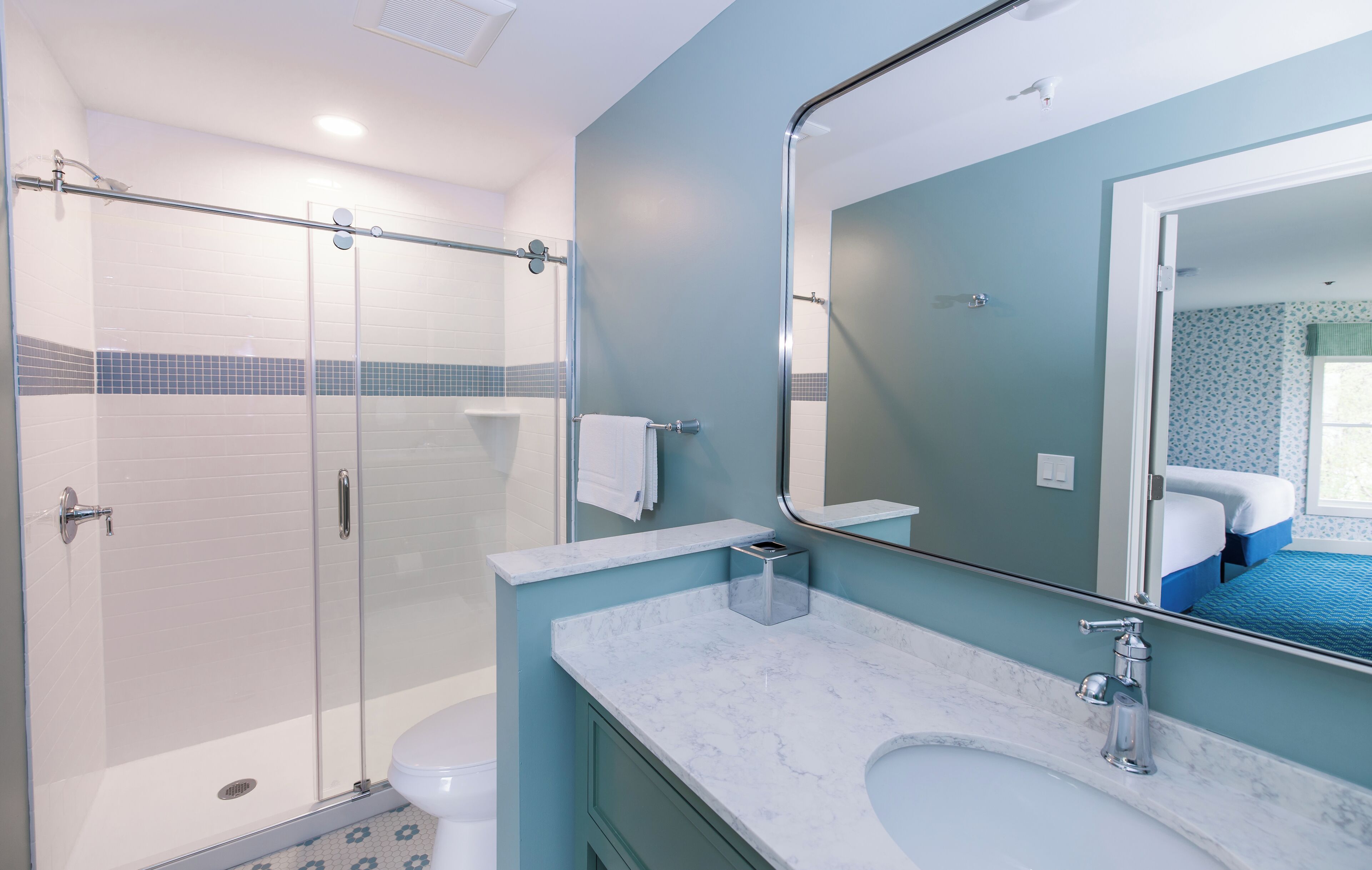 Une salle de bain moderne avec une douche à parois de verre, des toilettes blanches, un lavabo en marbre et un grand miroir qui reflète une chambre à coucher avec une couvre-lit bleue.