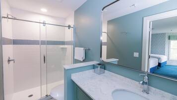 Une salle de bain moderne avec une douche à parois de verre, des toilettes blanches, un lavabo en marbre et un grand miroir qui reflète une chambre à coucher avec une couvre-lit bleue.