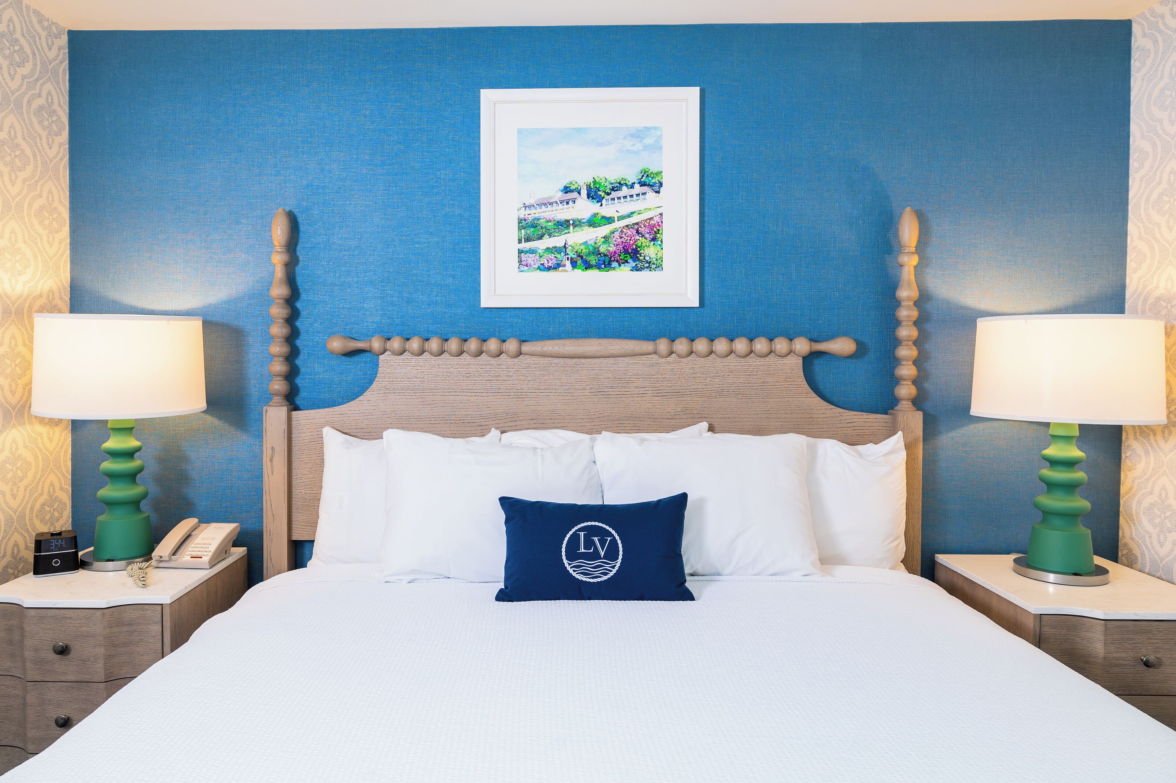 A hotel room with a wooden headboard, white bedding, a blue pillow with a logo, two bedside tables with lamps, and a framed picture on the wall.