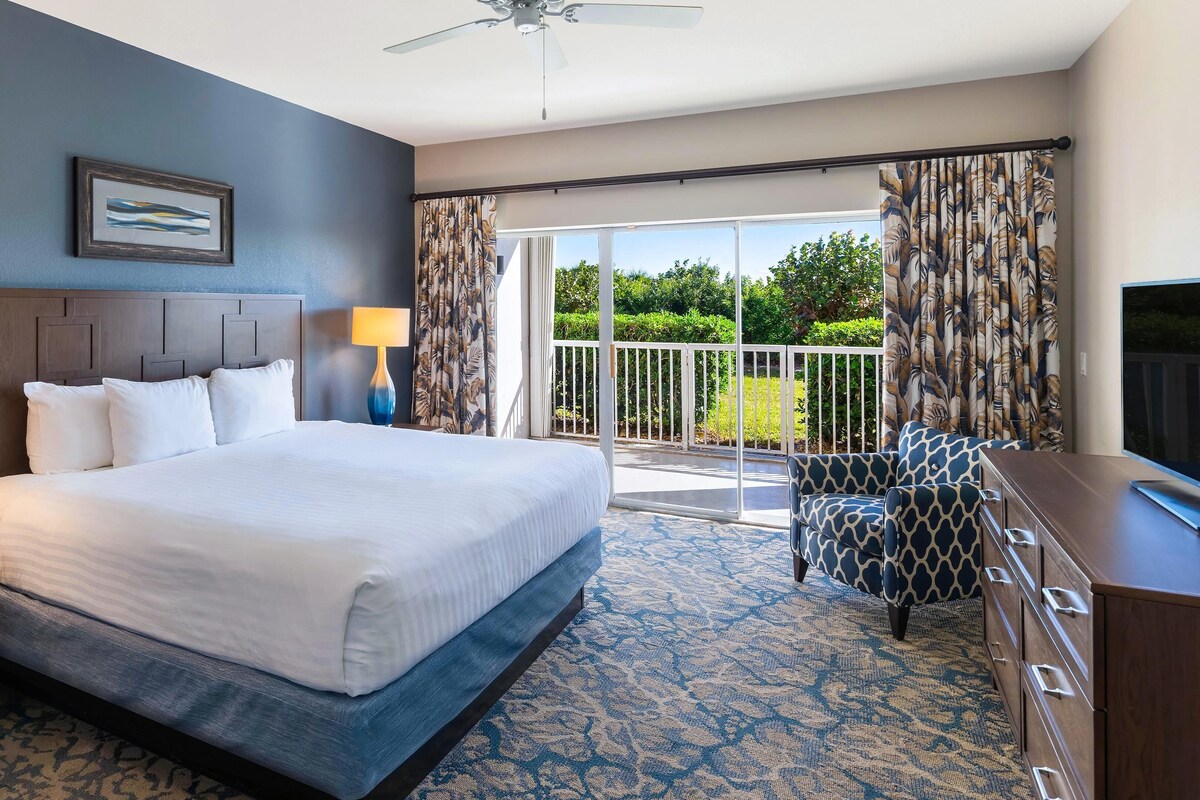 A hotel room with a bed, a desk, a TV, a ceiling fan, and a balcony with curtains.
