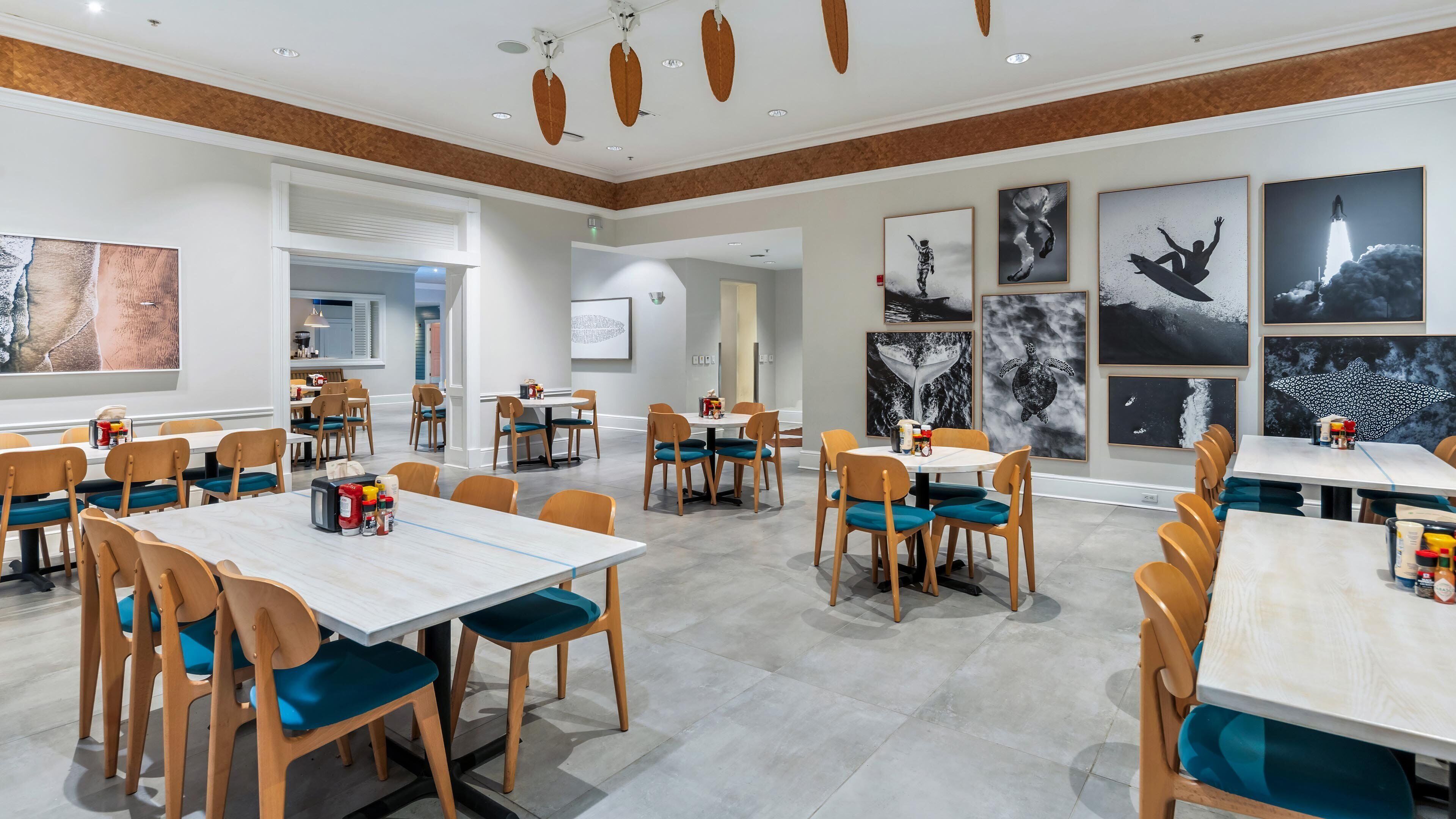 A spacious dining area with multiple tables and chairs, a kitchen in the background, and various framed artworks on the walls.
