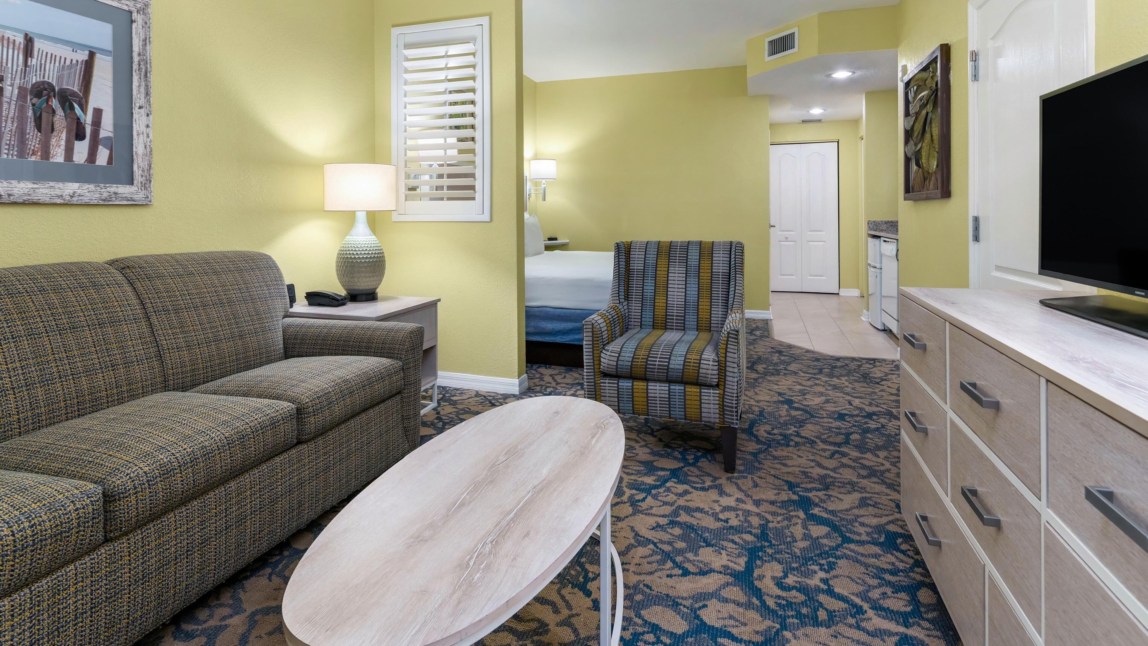 A hotel room with a sofa, armchair, small table, lamp, TV, and a door leading to another room.