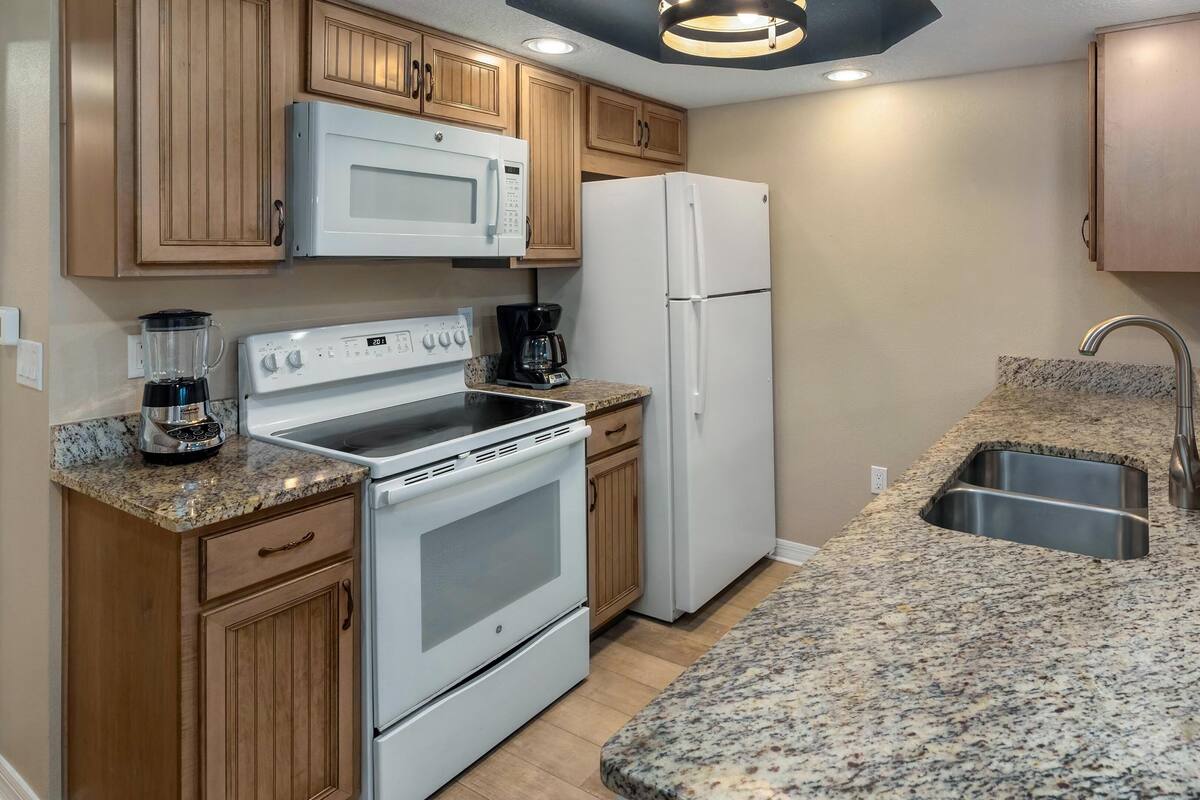 A kitchen with wooden cabinets, a white microwave, a white refrigerator, a stove, a coffee maker, and a granite countertop with a sink.